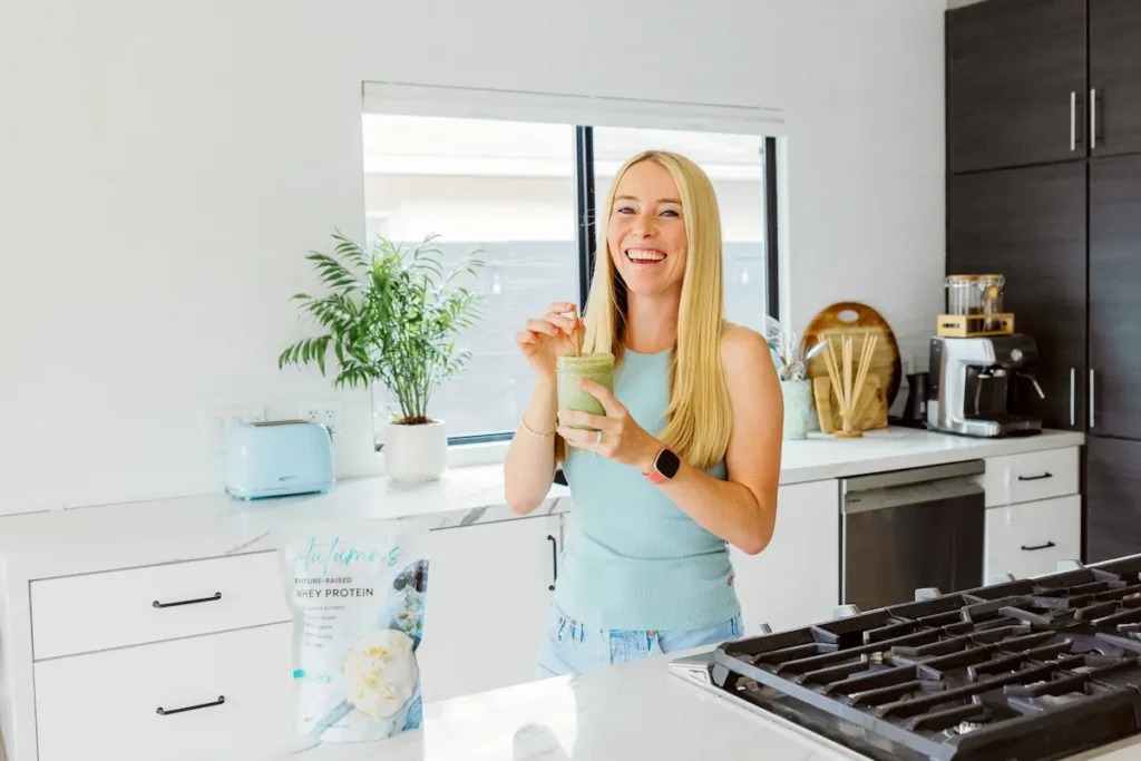 Autumn, author of Autumn Elle Nutrition, standing in kitchen drinking a protein shake and smiling.