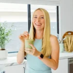 Autumn, author of Autumn Elle Nutrition, standing in kitchen drinking a protein shake and smiling.