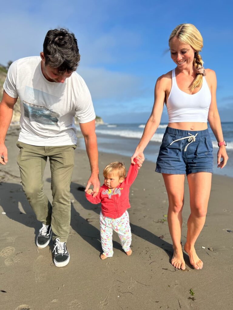 autumn bates and husband walking on the beach with a baby