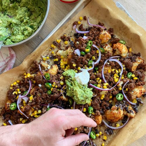 cauliflower nachos with guacamole, onions, ground beef, black beans and cheese with a hand reaching for one