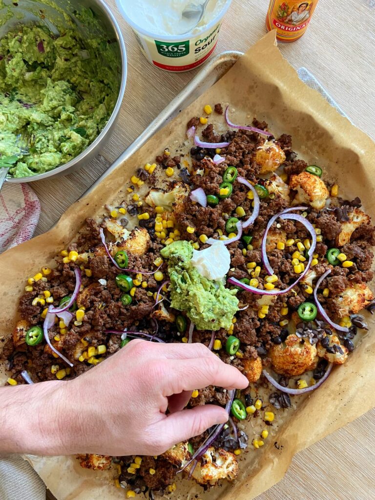 cauliflower nachos with guacamole, onions, ground beef, black beans and cheese with a hand reaching for one